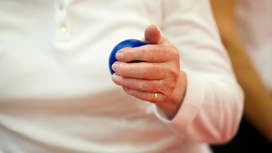 Nahaufnahme einer Hand, die einen blauen Ball hält, während die Person in einem weißen Oberteil sitzt.