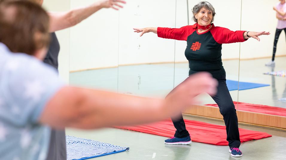 Eine ältere Frau in Sportkleidung führt eine Dehnübung in einem Fitnessraum mit Spiegeln aus.