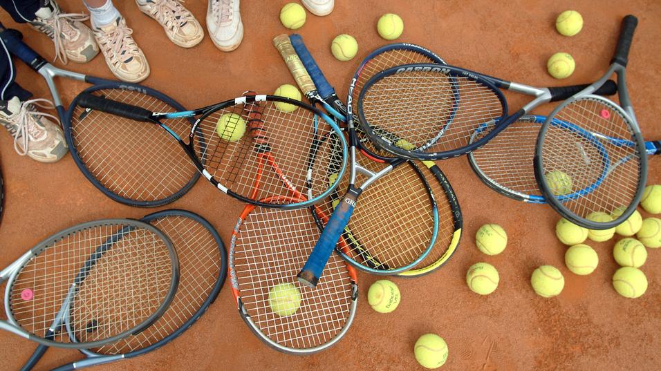 Ansammlung von Tennisschlägern und Bällen auf einem Sandplatz, umgeben von Füßen in Sportschuhen.