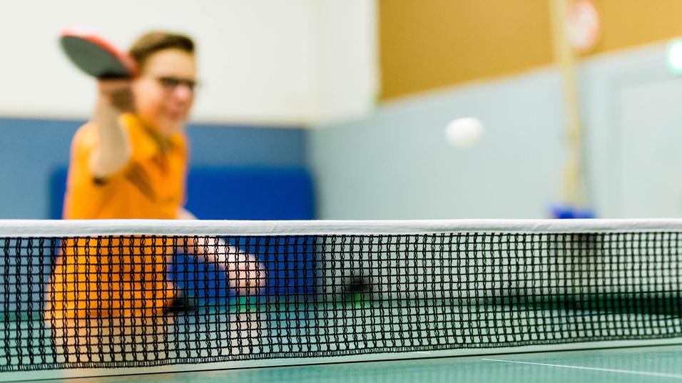 Tischtennisschläger in der Hand eines Spielers, im Hintergrund ein Ball über das Netz fliegend auf einer grünen Tischtennisplatte.