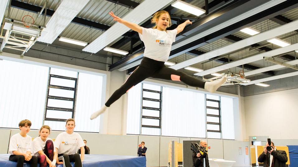 Mädchen springt mit ausgestreckten Armen über eine Trampolinfläche, während Zuschauer auf einer Bank sitzen.
