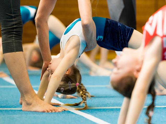 Trainer unterstützt ein Kind beim Rückwärtsspagat in einer Gymnastikhalle mit blauer Matte. Weitere Geräte im Hintergrund.