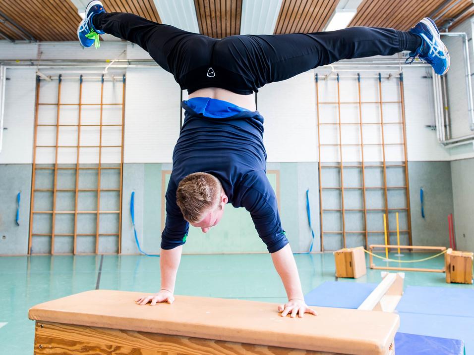 Junge macht Handstand auf einem Holztisch in einer Sporthalle mit verschiedenen Turngeräten im Hintergrund.