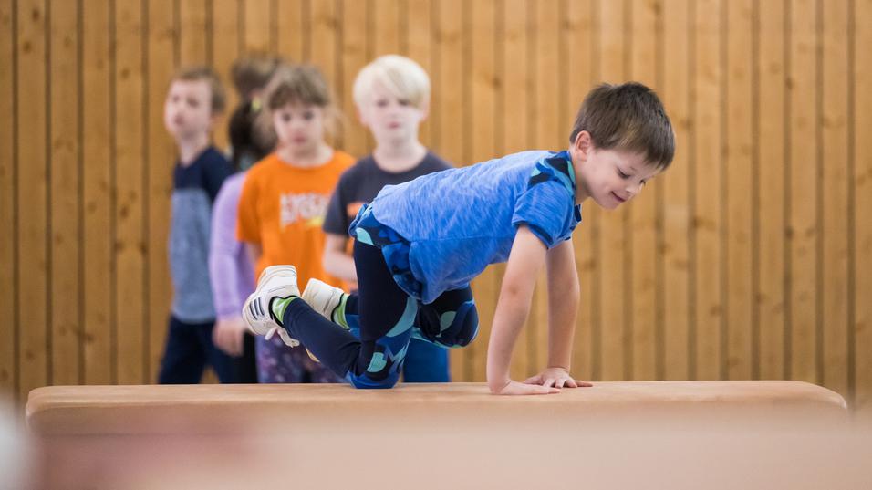 Ein Junge balanciert auf einem Blickkasten, während mehrere Kinder im Hintergrund zuschauen. Holzpaneele sind sichtbar.