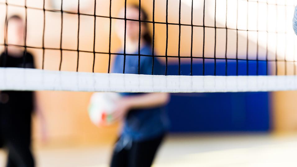 Unscharfe Silhouetten von zwei Personen hinter einem Volleyballnetz in einer Sporthalle. Eine Person hält einen Volleyball.